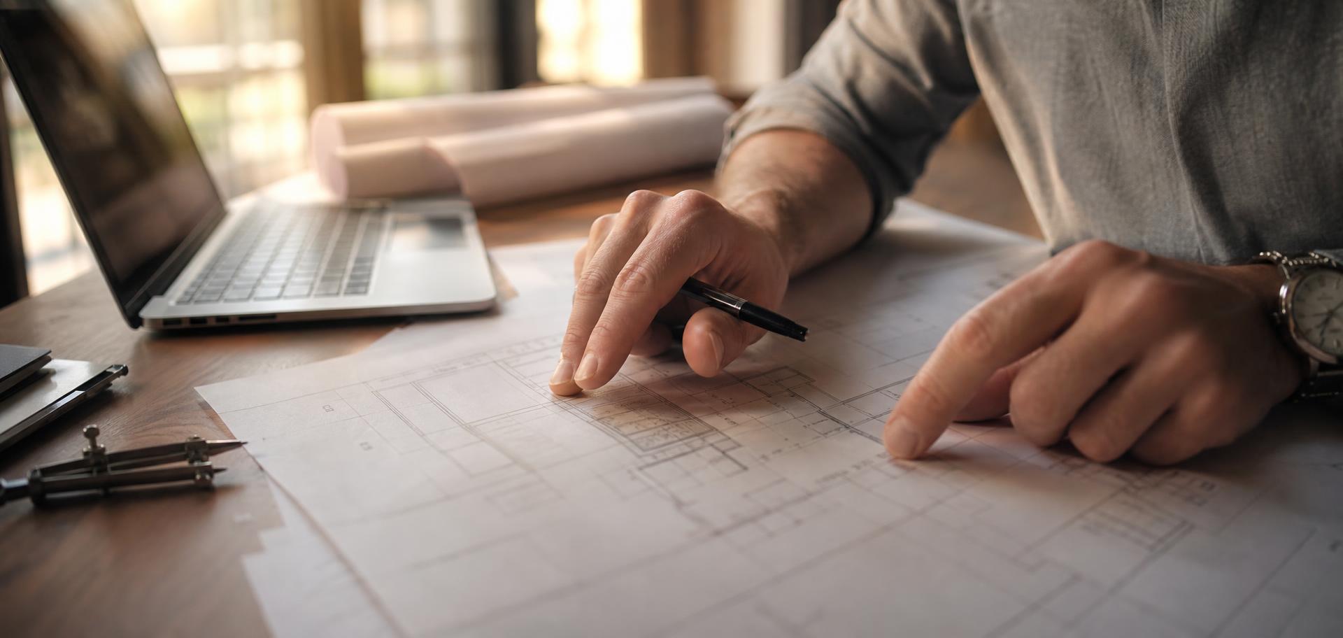 Close-up of hands sketching over architectural floor plan drawings on a wooden desk beside a laptop, a compass and a metal ruler in warm window light